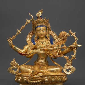 Pode incluir: Uma estátua dourada de uma divindade sentada com vários braços, adornada com uma coroa cravejada de joias e detalhes turquesa. A figura segura um arco e flecha e está sentada sobre uma base de flor de lótus. O texto "Nidhiratna" está na parte inferior.