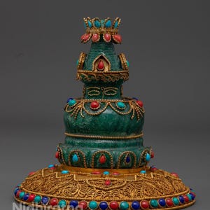 May include: An ornate, green, gold, and jewel-toned decorative object. The object has a tiered design with a wide base and a crown-like top. The base and top are adorned with red, blue, and turquoise gemstones. The object has intricate gold filigree details.