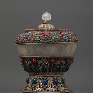 May include: An ornate, silver-toned vessel with a clear, spherical finial. The vessel is adorned with red, turquoise, and blue gemstones. The base and lid feature intricate metalwork. The overall design suggests a decorative or ceremonial object.