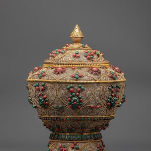 Può includere: Un vaso decorativo dorato ornato di pietre preziose rosse e verdi, con coperchio. Il vaso presenta intricati dettagli in filigrana e un terminale conico. Anche la base è dorata e decorata con pietre preziose. La parola "Nidhiratna" è in basso.