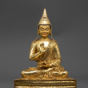 May include: A golden Buddha statue in a seated position, with one hand raised in a blessing gesture. The statue features intricate detailing on the robe and base, and a pointed hat. The statue is on a rectangular base with decorative patterns.