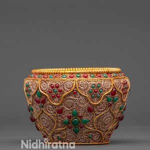 May include: Ornate, gold-toned metal bowl with intricate filigree and inlaid red and green gemstones. The bowl has a wide, decorative rim and a rounded body. The word "Nidhīratna" is printed on the side.