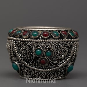 Peut inclure: Un bol argenté orné d'un motif floral détaillé. Le bol est orné de pierres précieuses rouges et turquoise alternées sur le bord et les côtés. Le texte "Nidhiratna" est visible sur le côté du bol.