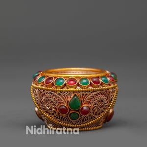 May include: A gold-coloured bowl with detailed filigree and a rim set with alternating red and green gemstones. The bowl has a central design of red and green gemstones. The brand name "Nidhiratna" is visible on the side.