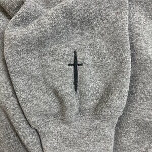 Op de afbeelding: Close-up van een grijze sweatshirtmouw met een zwart kruis geborduurd op de manchet. De stof heeft een getextureerde uitstraling. De manchet is geribbeld.
