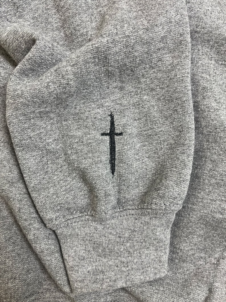 Op de afbeelding: Close-up van een grijze sweatshirtmouw met een zwart kruis geborduurd op de manchet. De stof heeft een getextureerde uitstraling. De manchet is geribbeld.