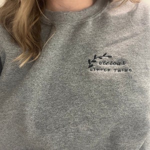 Op de afbeelding: Een grijze sweatshirt met de geborduurde tekst "vicious little thing" in zwart. De tekst wordt vergezeld door een klein zwart bladontwerp. De sweatshirt heeft een ronde hals.