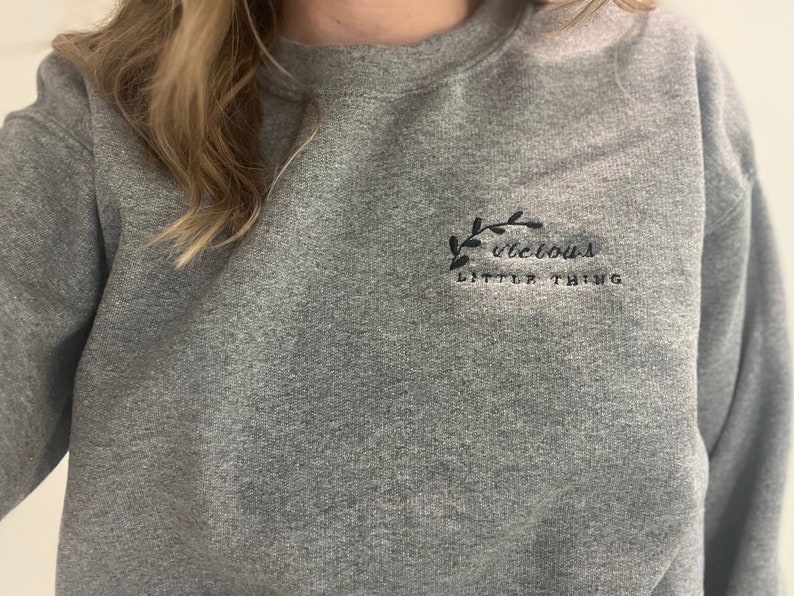 Op de afbeelding: Een grijze sweatshirt met de geborduurde tekst "vicious little thing" in zwart. De tekst wordt vergezeld door een klein zwart bladontwerp. De sweatshirt heeft een ronde hals.