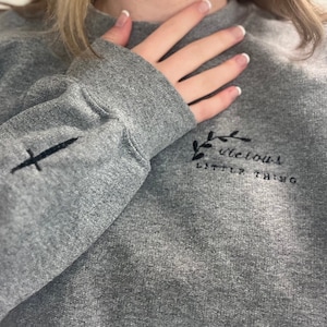 Op de afbeelding: Een grijze sweatshirt met geborduurde details. De linkermouw heeft een zwart zwaardontwerp, terwijl de borst de tekst "vicious little thing" met een bloemaccent toont. De sweatshirt heeft een ronde hals en lange mouwen.