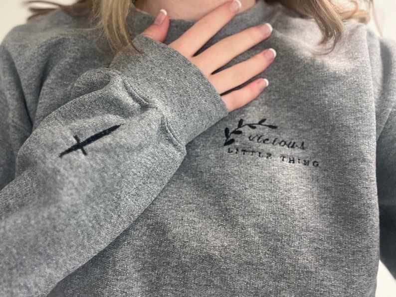 Op de afbeelding: Een grijze sweatshirt met geborduurde details. De linkermouw heeft een zwart zwaardontwerp, terwijl de borst de tekst "vicious little thing" met een bloemaccent toont. De sweatshirt heeft een ronde hals en lange mouwen.