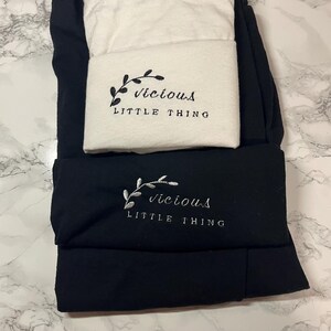 Op de afbeelding: Zwarte en witte leggings met geborduurde tekst en een botanisch ontwerp. De tekst luidt "Vicious Little Thing". De leggings zijn opgevouwen en op elkaar gestapeld, met het witte paar bovenop het zwarte paar.