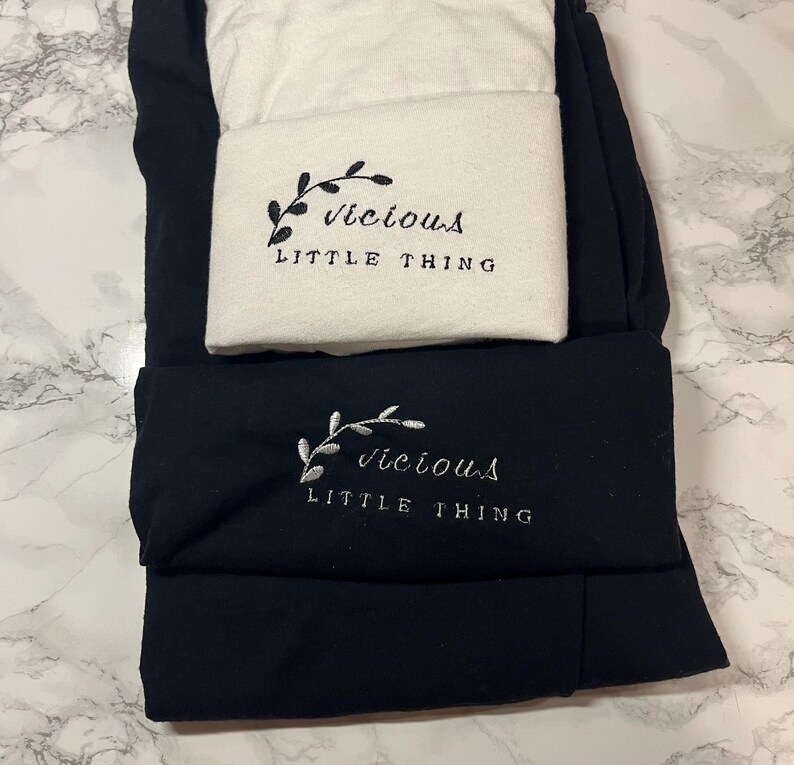 Op de afbeelding: Zwarte en witte leggings met geborduurde tekst en een botanisch ontwerp. De tekst luidt "Vicious Little Thing". De leggings zijn opgevouwen en op elkaar gestapeld, met het witte paar bovenop het zwarte paar.