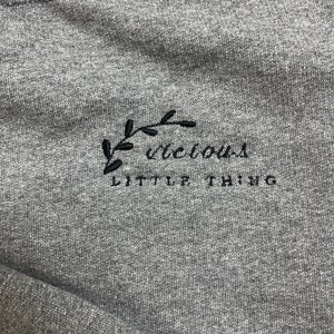 Op de afbeelding: Een grijze sweatshirt met de geborduurde tekst "Vicious Little Thing" in zwart. Een zwart bladontwerp staat boven het woord "Vicious". De sweatshirt is gemaakt van een zachte, getextureerde stof.
