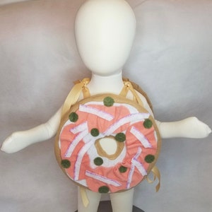 K&ouml;nnte beinhalten: Ein Donut-Kost&uuml;m aus Filz f&uuml;r Kinder. Der Donut ist braun mit rosa Zuckerguss und gr&uuml;nen Streuseln. Das Kost&uuml;m hat Tr&auml;ger, um es wie einen Rucksack zu tragen.