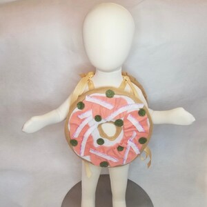 K&ouml;nnte beinhalten: Ein rosafarbenes und braunes Donut-Kost&uuml;m f&uuml;r ein Kind. Der Donut hat wei&szlig;en Zuckerguss und gr&uuml;ne Streusel. Das Kost&uuml;m hat Tr&auml;ger, um es wie einen Rucksack zu tragen.
