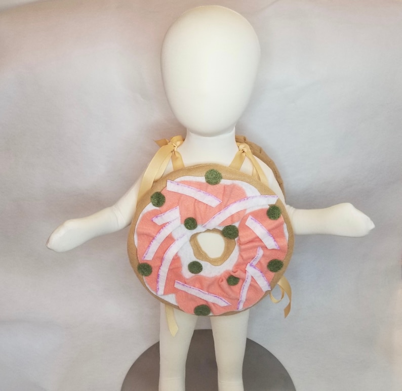 K&ouml;nnte beinhalten: Ein rosafarbenes und braunes Donut-Kost&uuml;m f&uuml;r ein Kind. Der Donut hat wei&szlig;en Zuckerguss und gr&uuml;ne Streusel. Das Kost&uuml;m hat Tr&auml;ger, um es wie einen Rucksack zu tragen.
