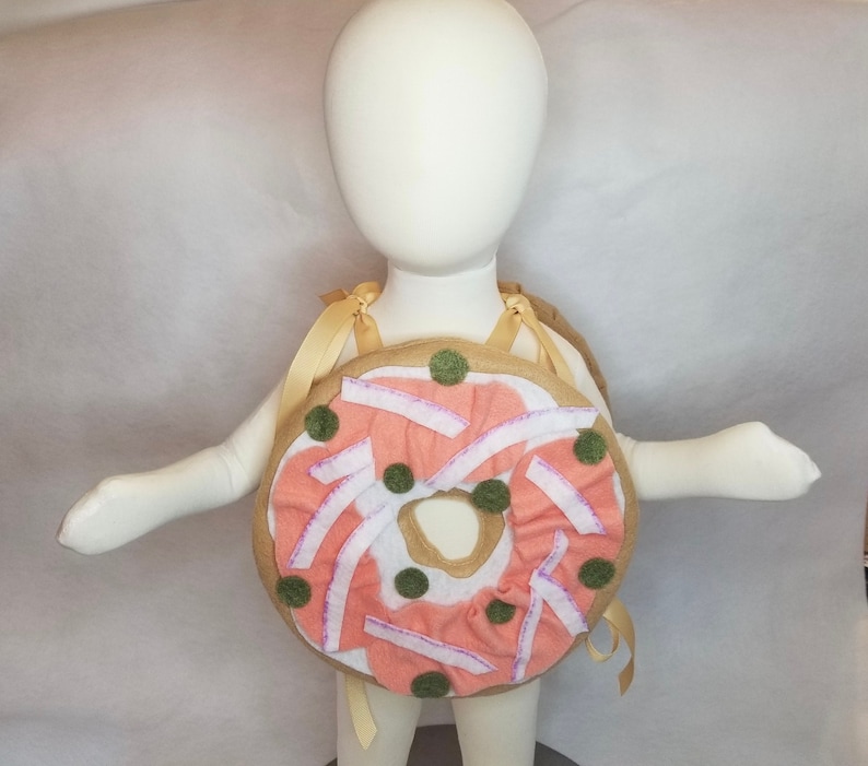 K&ouml;nnte beinhalten: Ein Donut-Kost&uuml;m aus Filz f&uuml;r Kinder. Der Donut ist braun mit rosa Zuckerguss und gr&uuml;nen Streuseln. Das Kost&uuml;m hat Tr&auml;ger, um es wie einen Rucksack zu tragen.
