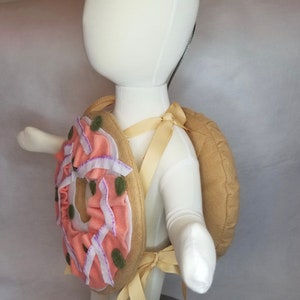 K&ouml;nnte beinhalten: Ein rosafarbener und gr&uuml;ner Donut-Rucksack mit einem braunen Filzr&uuml;ckenpanel und verstellbaren Tr&auml;gern. Der Donut hat rosa Zuckerguss und gr&uuml;ne Streusel.