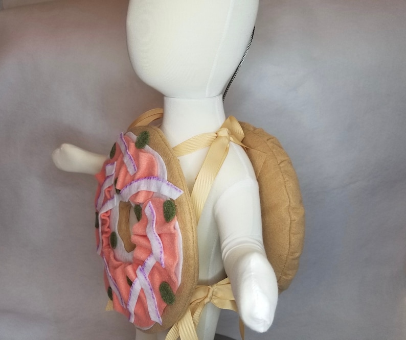 K&ouml;nnte beinhalten: Ein rosafarbener und gr&uuml;ner Donut-Rucksack mit einem braunen Filzr&uuml;ckenpanel und verstellbaren Tr&auml;gern. Der Donut hat rosa Zuckerguss und gr&uuml;ne Streusel.
