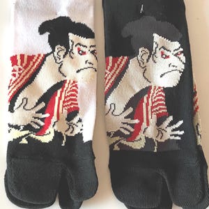 May include: Pair of tabi socks, one white and one black, each featuring a graphic of a stern-faced Japanese figure. The socks have a split-toe design and red, black, and white accents.