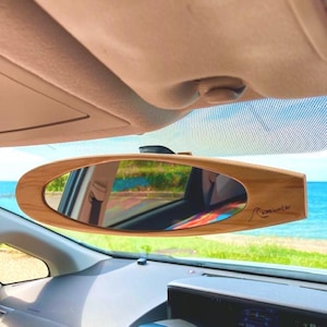 May include: A surfboard-shaped wooden rearview mirror, mounted inside a car. The mirror reflects a view of the ocean and a car interior. The text "wave hunter" is printed on the right side of the mirror.