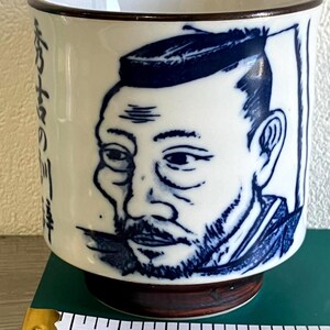 Puede incluir: Taza de té de cerámica blanca con una ilustración azul y blanca de la cara de un hombre. La taza tiene un borde y una base marrones. Texto japonés visible en el lateral de la taza.