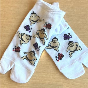 Op de afbeelding: Witte tabi sokken met illustraties van sumoworstelaars in bruin en zwart. De sokken hebben ook kleine paarse en rode bloemmotieven. De sokken zijn gemaakt van een zacht, geribbeld materiaal.