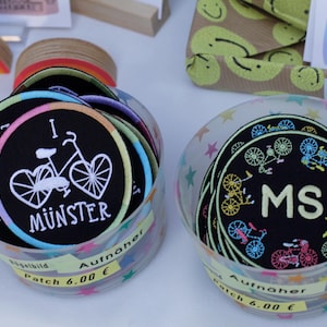 Könnte beinhalten: Zwei durchsichtige Plastikbehälter gefüllt mit bunten gestickten Aufnähern. Ein Behälter enthält Aufnäher mit einem Fahrrad-Design und dem Text "I ♥ MÜNSTER". Der andere Behälter enthält Aufnäher mit einem Stern-Design und dem Text "MS".