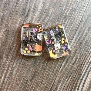 Pode incluir: Dois chaveiros retangulares de resina com designs de Halloween. Um apresenta a letra "R" e as iniciais "JS", uma caveira e uma abóbora. O outro tem a letra "L" e as iniciais "JS", um morcego e um fantasma.