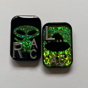 May include: Two black and green square-shaped pins with silver lettering. The first pin features a green alien with the letters "RAC" on the right side. The second pin features a black UFO with the letters "LAC" on the right side.