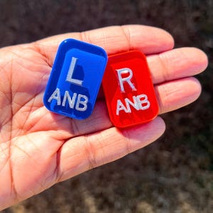 May include: Two rectangular plastic markers with rounded corners. One marker is blue with the white embossed letters L and ANB. The other marker is red with the white embossed letters R and ANB. Both markers feature a smooth, glossy finish.