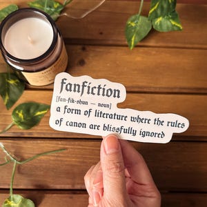 May include: A white sticker with black text that reads "fanfiction [fan-fik-shun - noun] a form of literature where the rules of canon are blissfully ignored."