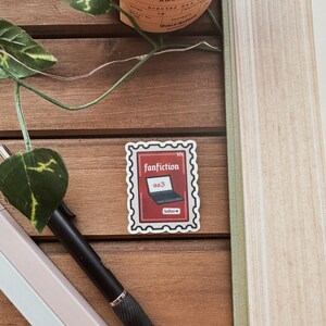 May include: A red and white sticker featuring a laptop with the text "ao3" on the screen. The sticker reads "fanfiction" and "hobos". The sticker is placed on a wooden surface.