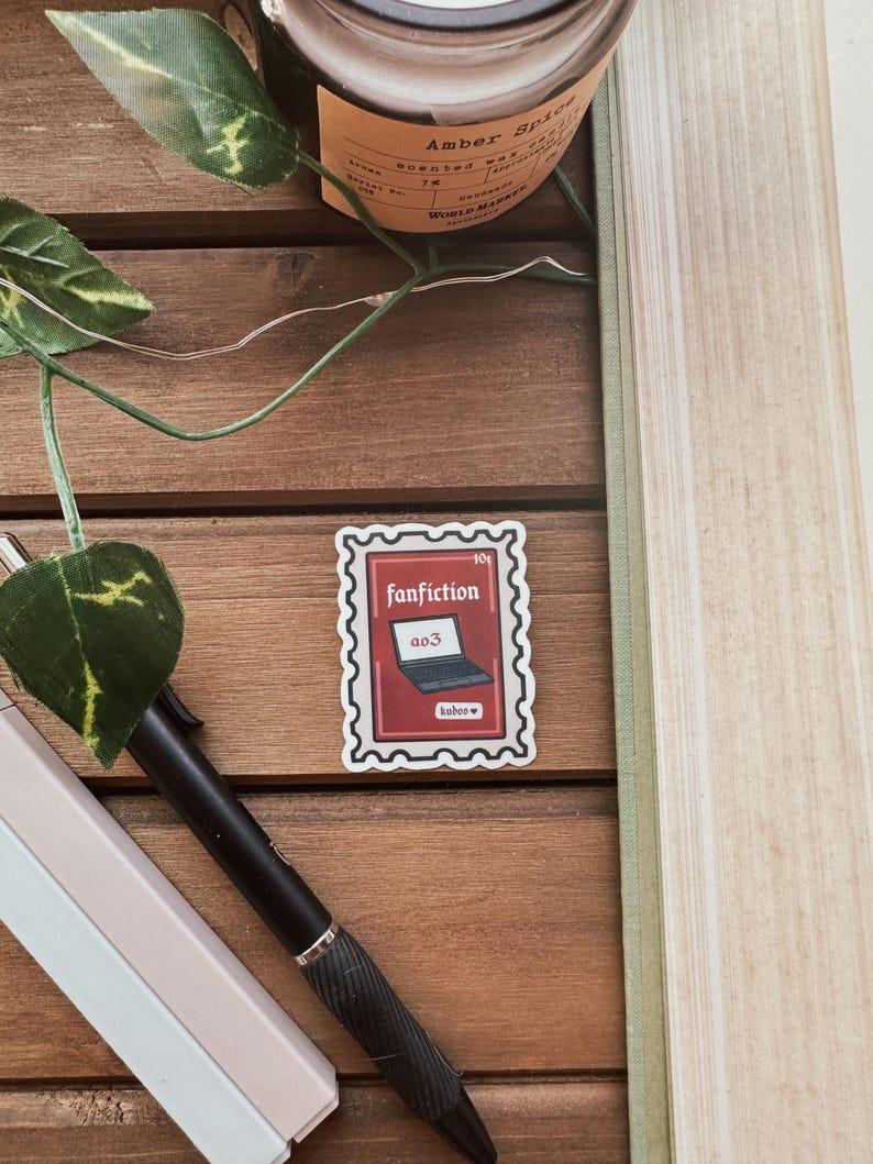 May include: A red and white sticker featuring a laptop with the text "ao3" on the screen. The sticker reads "fanfiction" and "hobos". The sticker is placed on a wooden surface.