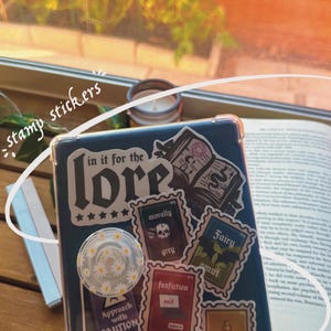 May include: A blue phone case with various stickers, including a large sticker that says "in it for the lore" with five stars underneath. Other stickers include "morally grey", "fanfiction", "fairy smut", and "approach with caution".