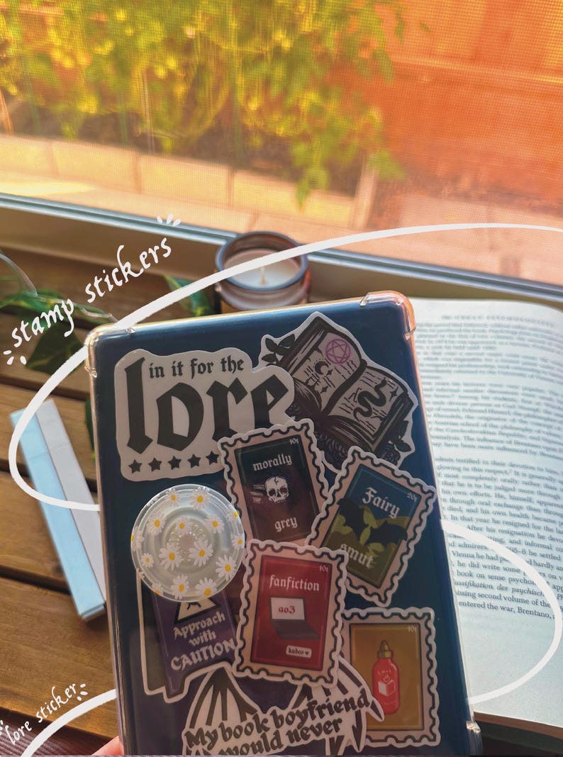 May include: A blue phone case with various stickers, including a large sticker that says "in it for the lore" with five stars underneath. Other stickers include "morally grey", "fanfiction", "fairy smut", and "approach with caution".