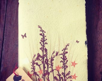 Peut inclure: Un journal artisanal sans arbre avec un motif floral sur la couverture. Le journal est fabriqué à partir de papier recyclé et porte une étiquette en papier brun sur laquelle est inscrit "Tree Free Handmade Paper Journal - 40 pages www.pulppaperpaper.com".