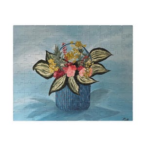 May include: A completed jigsaw puzzle featuring a still life of flowers in a blue basket. The flowers are a mix of pink, yellow, and red, with large green leaves. The background is a light blue.