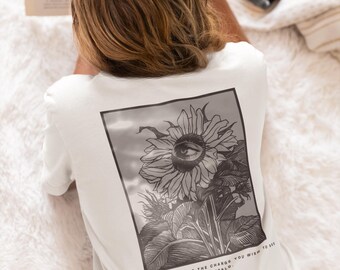 May include: White t-shirt featuring a black and white sunflower graphic with an eye at its centre. The text "YOU MUST BECOME THE CHANGE YOU WISH TO SEE IN THE WORLD" is printed below. The shirt is worn by a person, lying down.