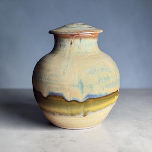 Op de afbeelding: Een keramieken pot met deksel, voorzien van een ronde vorm en een smalle hals. Het glazuur vertoont een mix van crème, lichtblauwe en saliegroene tinten, met een donker olijfgroene en bruine band rond het onderste deel van de pot.