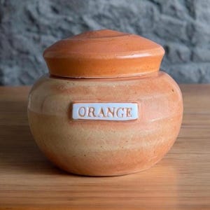 May include: A small, orange ceramic jar with a lid and a white label that says "ORANGE".