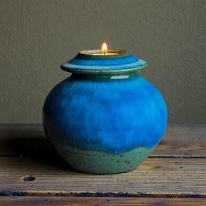 Op de afbeelding: Een blauwe keramische kandelaar met een brandende kaars. De kandelaar heeft een ronde body met een uitlopende bovenkant en een groene basis. De kaarsvlam is helder tegen het blauw en groen geglazuurde keramiek.