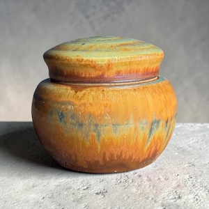 Op de afbeelding: Een ronde keramische pot met bijpassend deksel, voorzien van een gevarieerd glazuur in tinten warm oranje, diepbruin en gedempt blauwgroen. Het oppervlak heeft een gladde, glanzende afwerking met verticale druippatronen en een bolvormig lichaam.