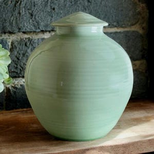 Puede incluir: Una urna de cerámica de color verde claro con tapa. La urna tiene una forma redonda y lisa y un acabado brillante.