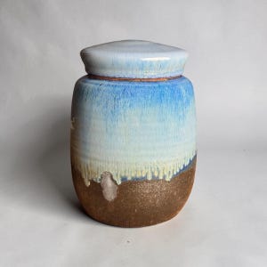 Peut inclure: Un pot en céramique avec couvercle, doté d'un corps cylindrique passant d'une partie supérieure émaillée lisse bleu clair et blanc à une base texturée brun foncé non émaillée, avec des coulures visibles là où l'émail rencontre l'argile.