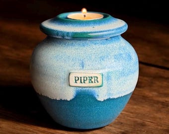 May include: A blue and white ceramic candle holder with a lit tea light on top. The candle holder has a lid and a label that reads "PIPER". The candle holder is on a wooden surface.