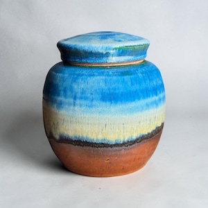 Op de afbeelding: Een keramische pot met deksel voorzien van een verloopglazuur dat overgaat van een diep terracotta bruin aan de basis naar lichtgeel, vervolgens helder hemelsblauw en tot slot een donkerder blauw op het deksel, met zichtbare druppelpatronen.