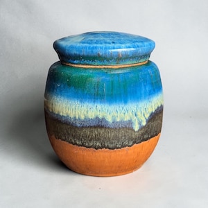 Puede incluir: Un tarro de cerámica con tapa que presenta un esmalte multicolor. La tapa y la parte superior son de un azul vibrante, que transiciona a una banda verde, un patrón de goteo amarillo pálido, una sección central marrón oscuro y una base naranja terracota.
