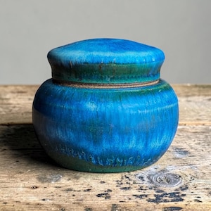 Op de afbeelding: Een ronde keramische pot met deksel, voorzien van een levendige blauwe en diepgroene glazuurlaag. Het oppervlak heeft een glanzende, meerkleurige uitstraling met subtiele verticale kleurstrepen die van boven naar de basis van de pot lopen.