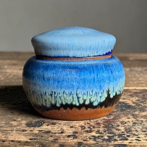 May include: A small ceramic jar with a lid, featuring a rounded shape and a reactive glaze that transitions from a light blue top to a vibrant blue middle, fading into a pale green drip pattern over a solid, unglazed brown clay base.